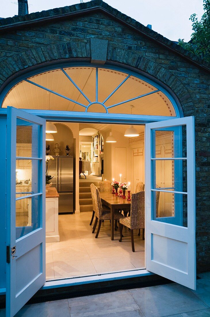 Open double glass doors and segmented transom window in brick facade; view into kitchen-dining room in romantic candlelight
