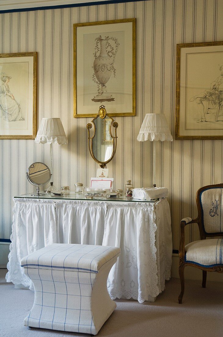 Mirror and pots on dressing table with glass top and lace valance below antique drawings on striped wallpaper