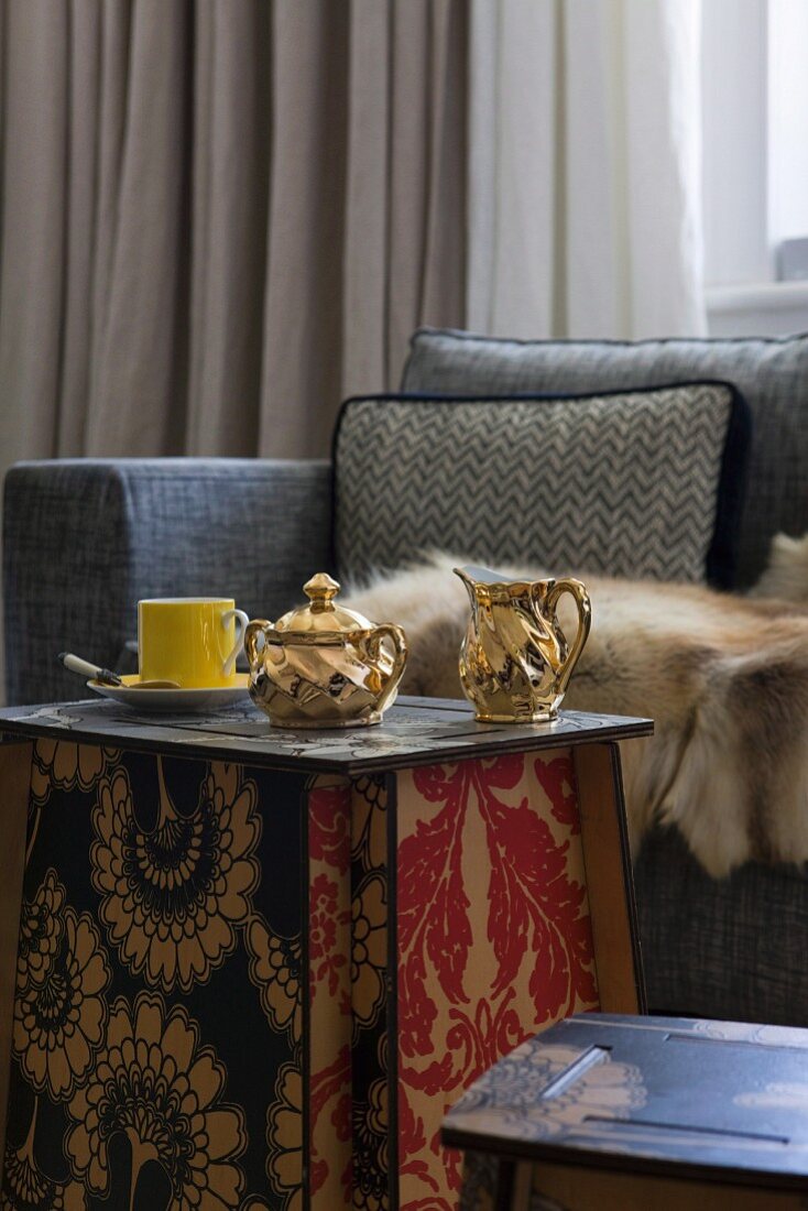 Milchkanne und Zuckerdose aus vergoldetem Porzellan auf Beistelltisch aus Steckplatten mit floralem Jugendstil-Muster; Tierfell auf Sofa im Hintergrund
