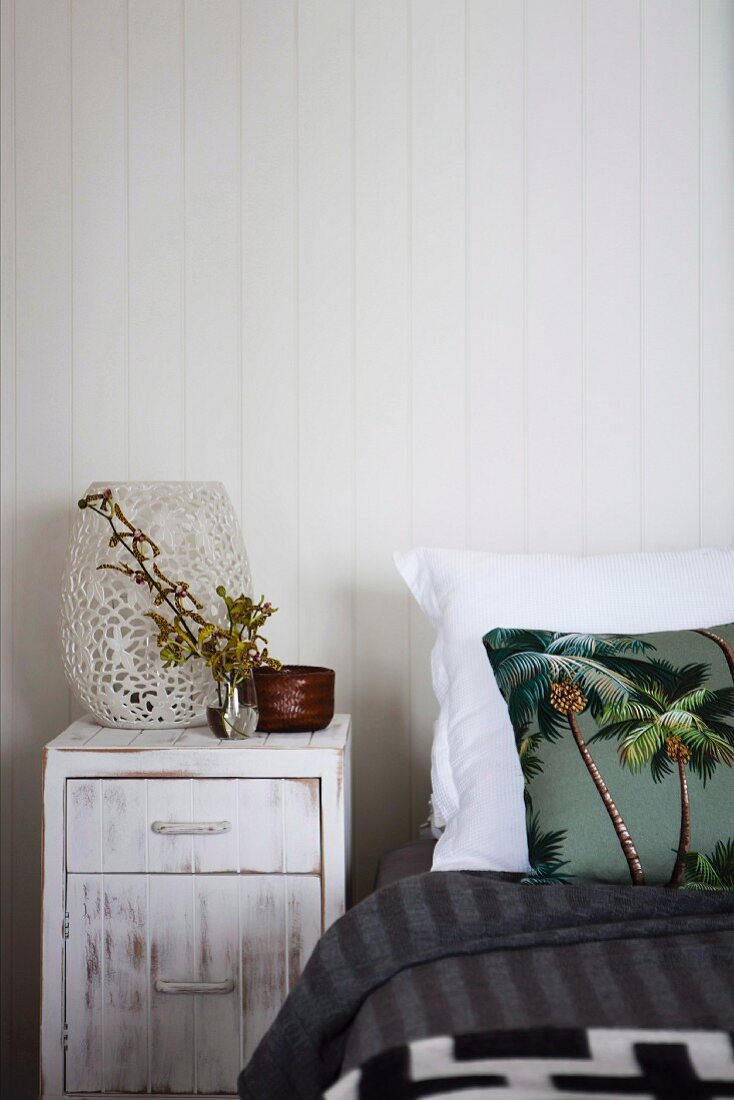 Weisses Nachtkästchen im Shabby Stil, neben teilweise sichtbarem Bett mit Kissen, darauf Palmenmotiv