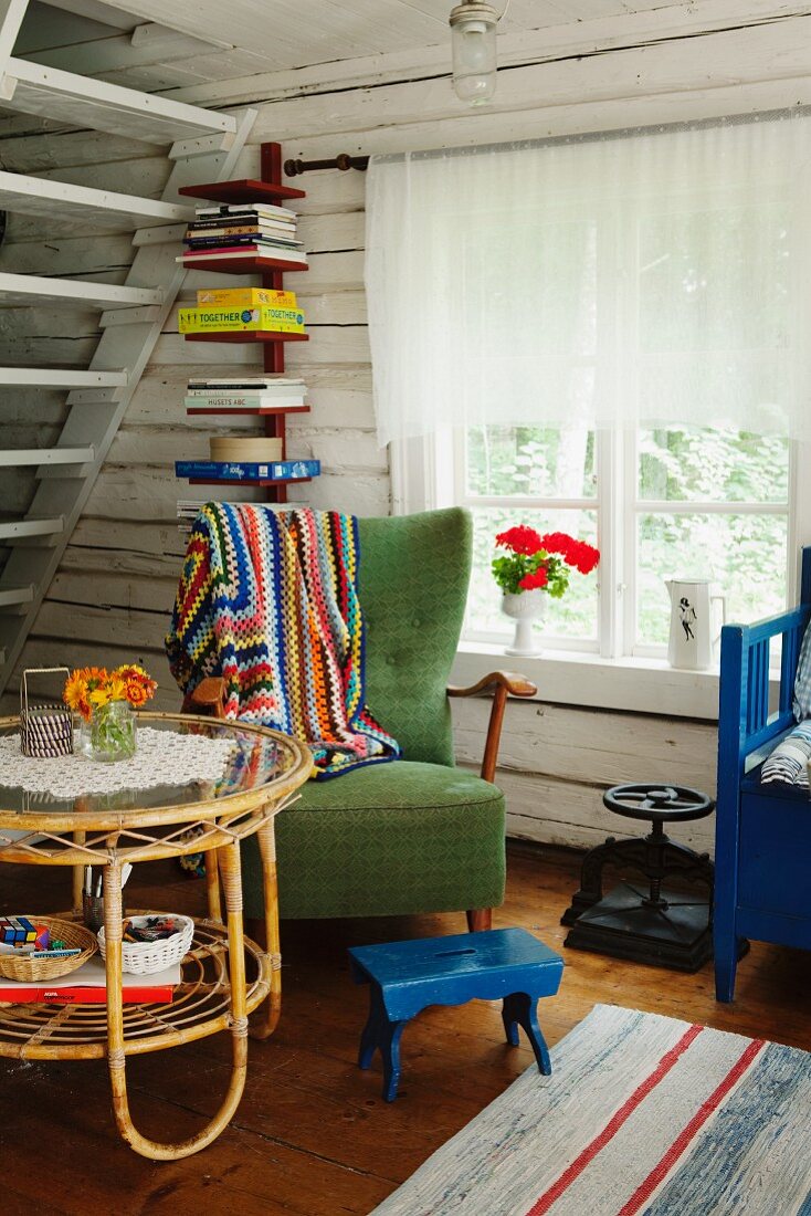 Rattan-Beistelltisch und grüner Sessel mit blauem Fussschemel in rustikalem Wohnraum eines Holzhauses