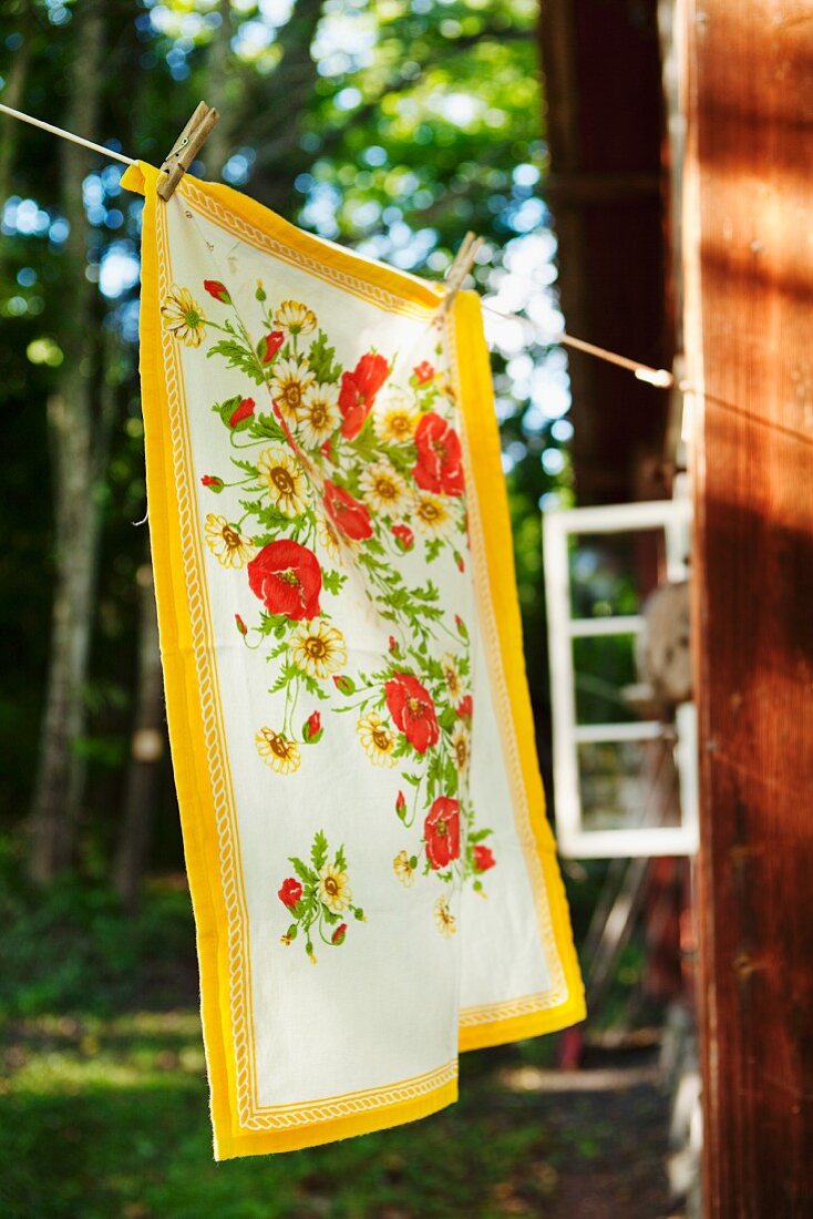 Geschirrtuch mit Blumenmuster an Wäscheleine aufgehängt vor Holzhäuschen im Wald