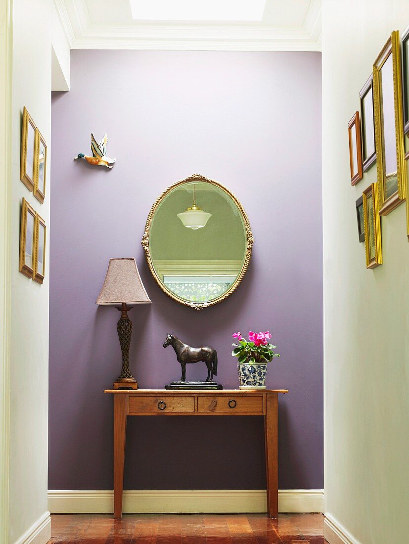 Horse figurine and table lamp on antique console table and mirror on lilac-painted wall in hallway