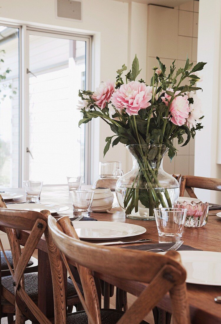 Glass vase of peonies on set table; detail of rustic, wooden chairs in foreground