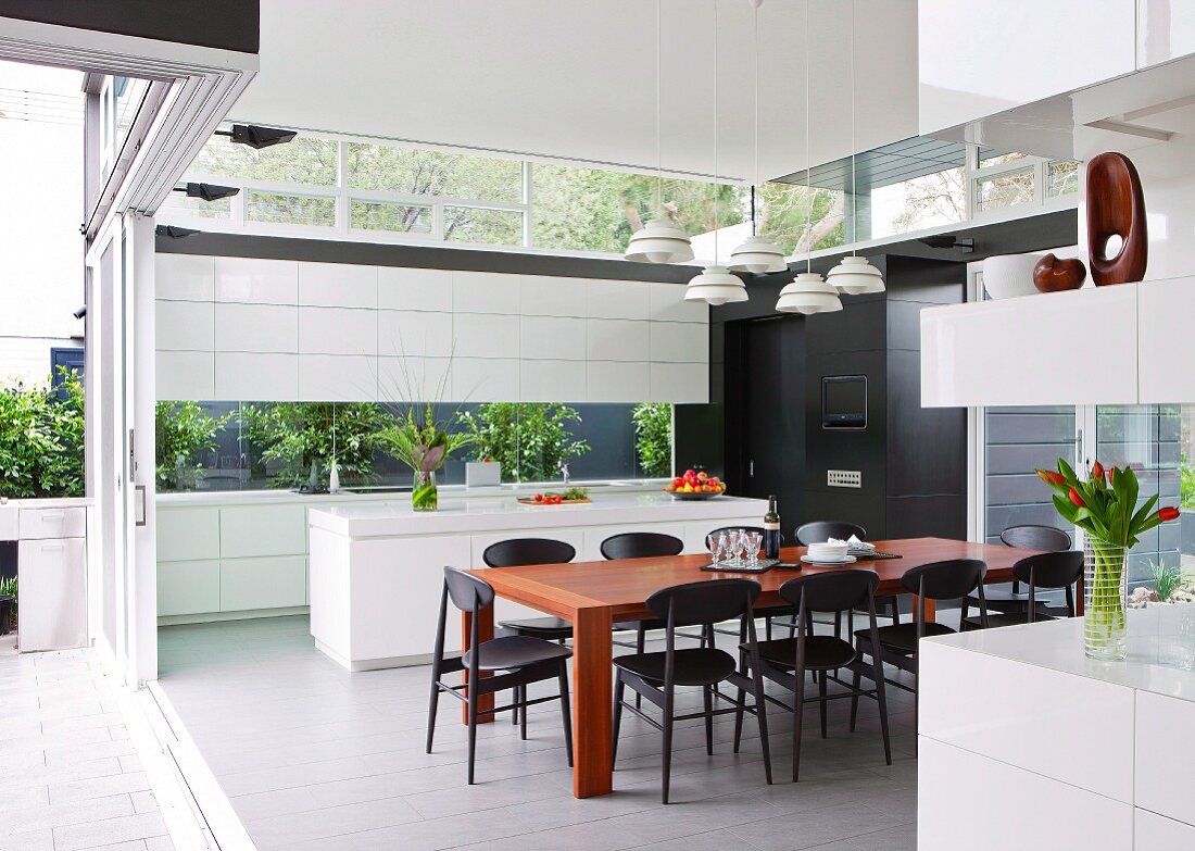 White, designer kitchen with wooden dining table and delicate chairs; open sliding glass wall leading to terrace on one side