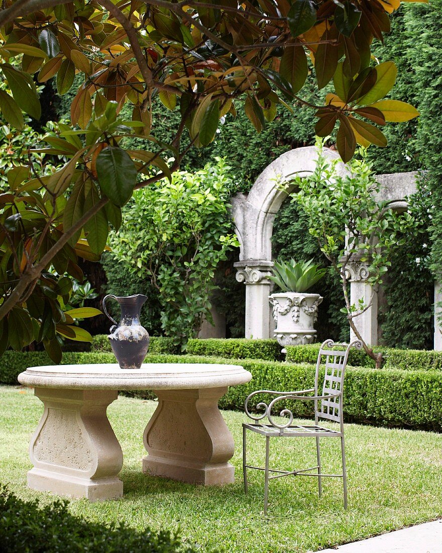 Metallstuhl an klassischem Steintisch im Garten; antike Säulen mit Rundbogen und Amphore hinter niedriger Buchsbaumhecke