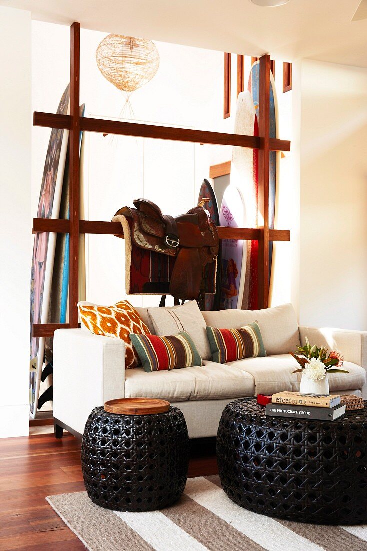 Pale sofa and black, wicker side tables; horse saddle on wooden, frame-like partition and surfboards in background