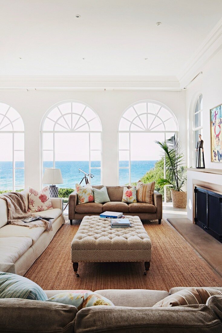 Helle Sofagarnitur und gepolsterter Couchtisch in mediterranem Wohnzimmer mit Blick durch Rundbogen Fenstertüren auf das Meer