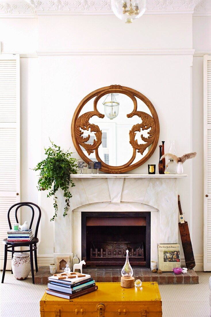 Partially visible, yellow wooden trunk in front of open fireplace and round mirror with carved wooden frame on mantelpiece