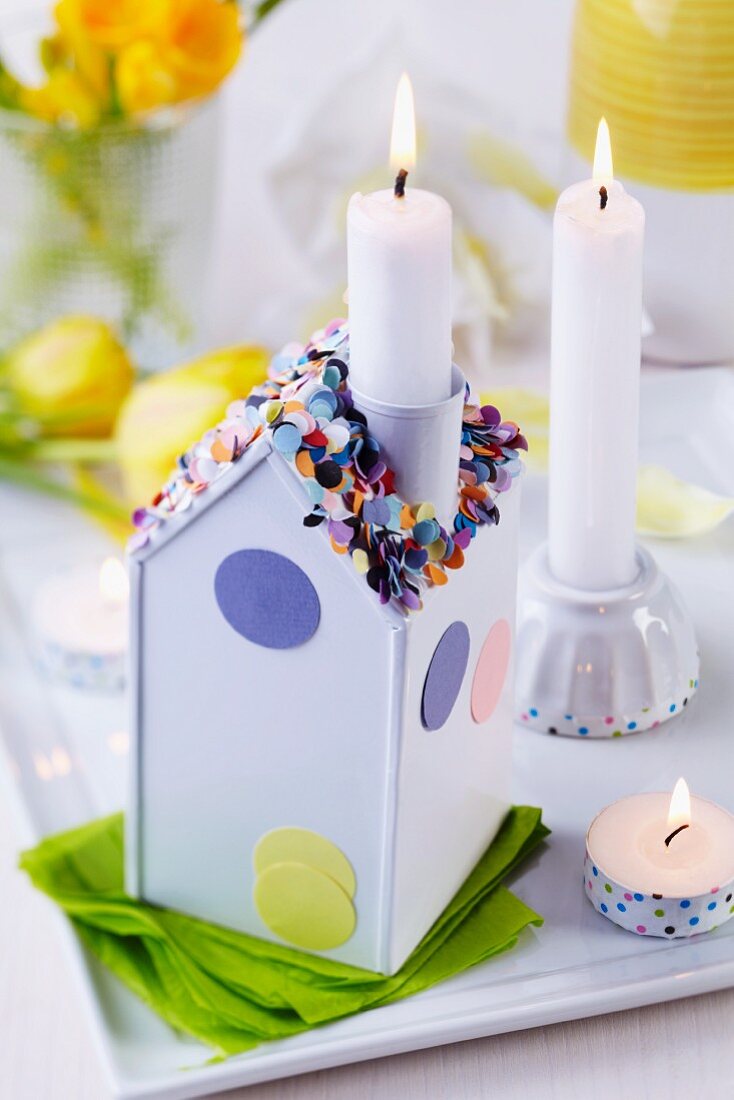 Lit candle in chimney of house-shaped candlestick decorated with confetti