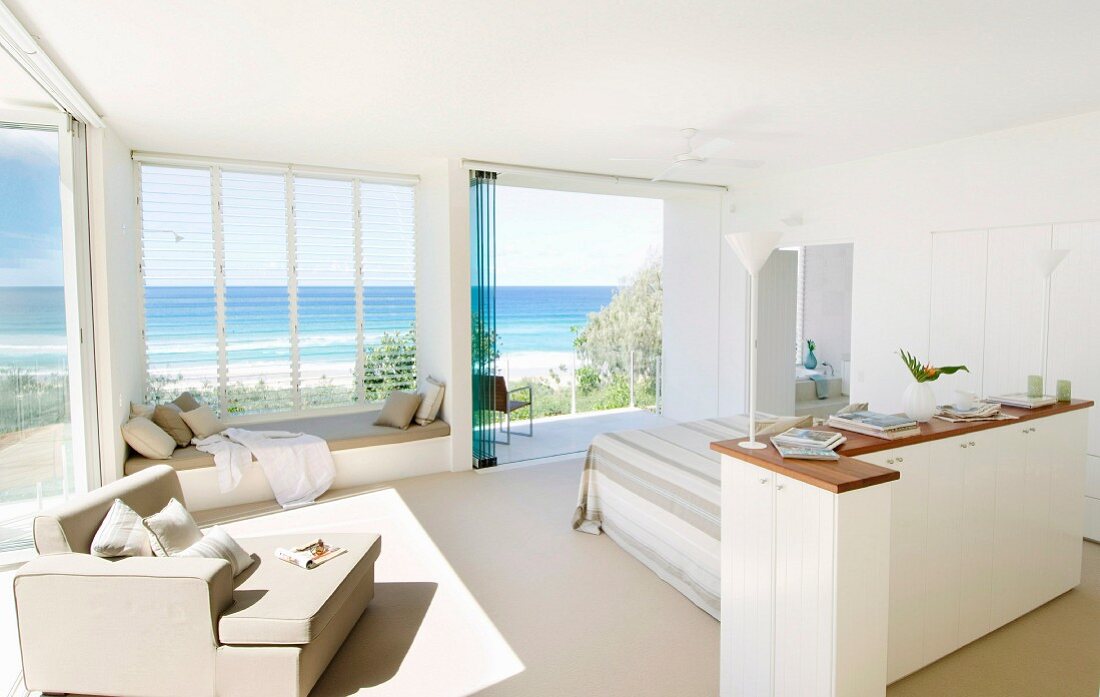 Grosszügiger, lichtdurchfluteter Schlafraum in Weiß, seitlich halbhoher Schrank vor Bett, Sessel vor Fenster mit Panoramablick auf das Meer