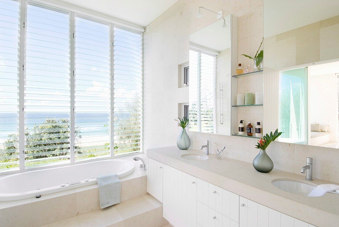 Moderne Waschzeile mit Steinplatte auf weissen Unterschränken, Stufe vor eingebauter Badewanne am Fenster mit Panoramablick