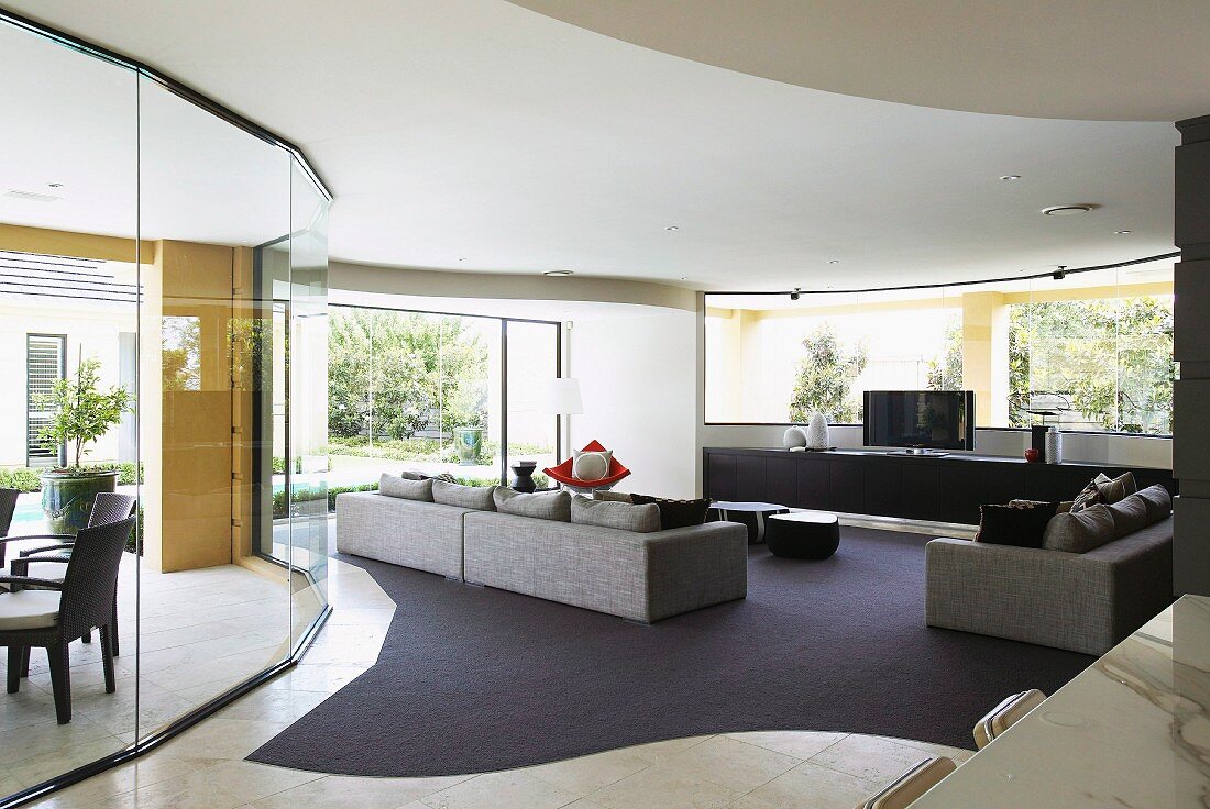 Grossräumiges, modernes Wohnzimmer, kubische Sofagarnitur auf Teppichboden mit geschwungenen Formen, seitlich gebogene Glas Trennwand