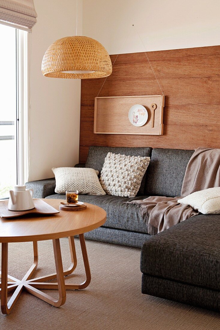 Pale wooden coffee table, grey corner sofa and place setting on tray hung on wooden wall in comfortable seating area