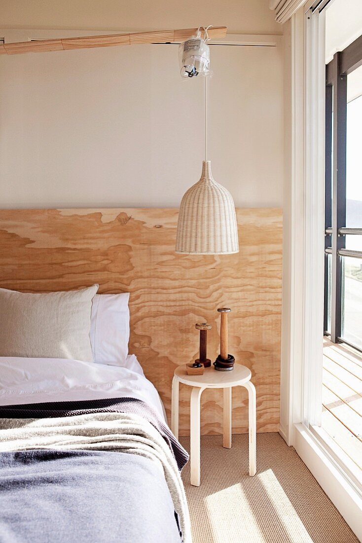 Sonnige Schlafzimmerecke, Pendelleuchte mit hellem Rattanschirm über Beistelltisch neben teilweise sichtbarem Bett, an Wand rustikale Holzplatte