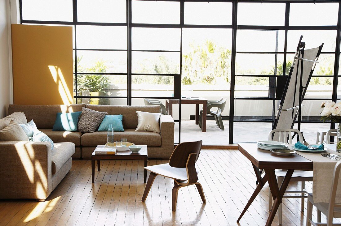 Wohn-Essbereich mit Stahl-Glasfassade vor Terrasse mit Ausblick; naturfarbene Sofakombination mit hellblauen Kissen und Klassiker Holzstuhl, daneben gedeckter Esstisch
