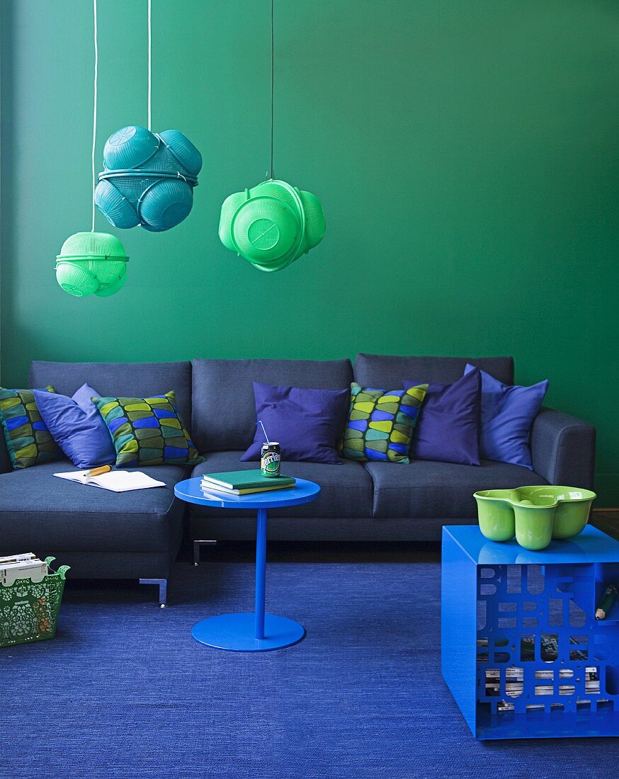 Blau und Grün sind die Farben der Wahl - blaues Polstersofa mit Kissen an grüner Wand, Pendellleuchten aus lackierten Küchensieben, blauer Hocker mit grüner Schale