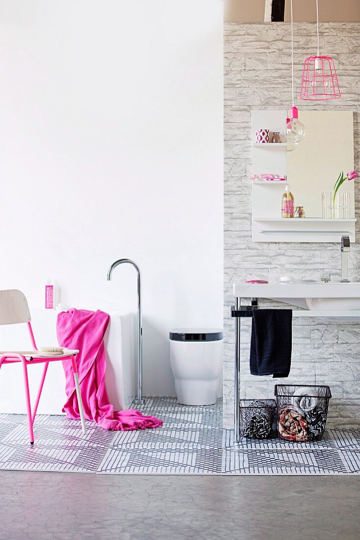 Badezimmer mit minimalistischem Waschtisch, darunter Körbe, an der Seite Stuhl mit pinkfarbenem Metallgestell, Handtuch in kräftigem Pink auf Badewanne mit Standarmatur