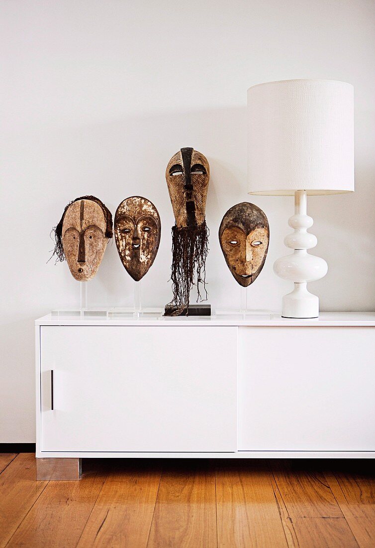 Afrikanische Masken auf transparenten Kunststoff Stelen neben weisser Tischleuchte auf Designer-Sideboard