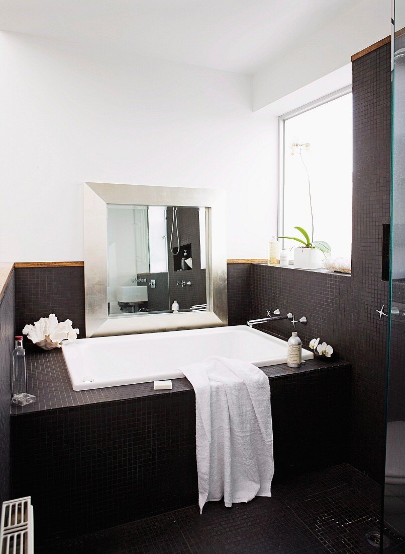 Bathtub in niche built into niche with window, silver-framed mirror leaning against wall, black mosaic tiled on bath surround and walls