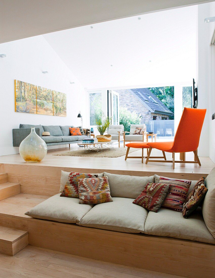 Seat cushions on broad step used as bench in front of modern lounge area on platform with sofa & orange chair