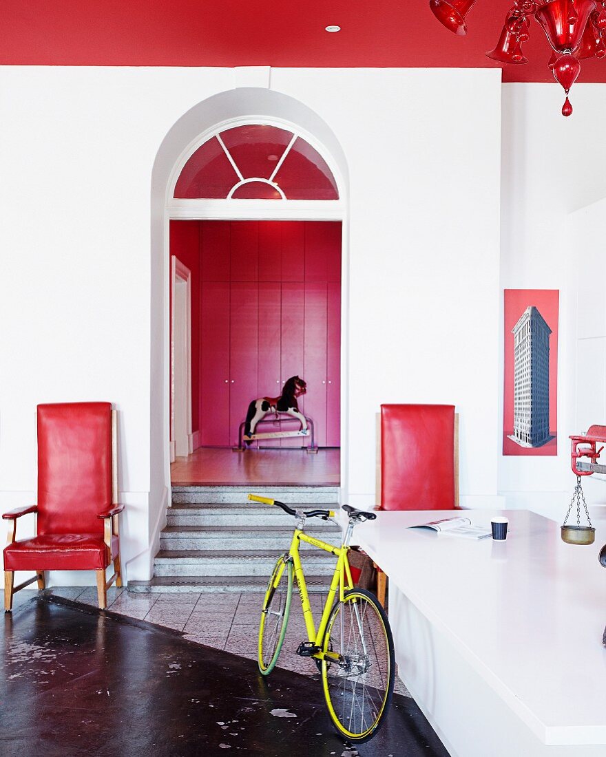 Bicycle leaning against counter opposite doorway with steps flanked by red armchairs; rocking horse in front of red fitted cupboards in background