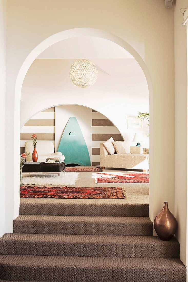 Treppenaufgang mit Teppichbelag vor Rundbogen und Blick in Wohnbereich mit folkloristischen Teppichen und modernen Möbeln