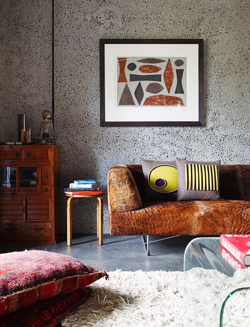 Bodenkissen auf flokatiartigem Teppich und Sofa mit Kuhfellbezug vor grauer Paneelwand mit gerahmtem Bild