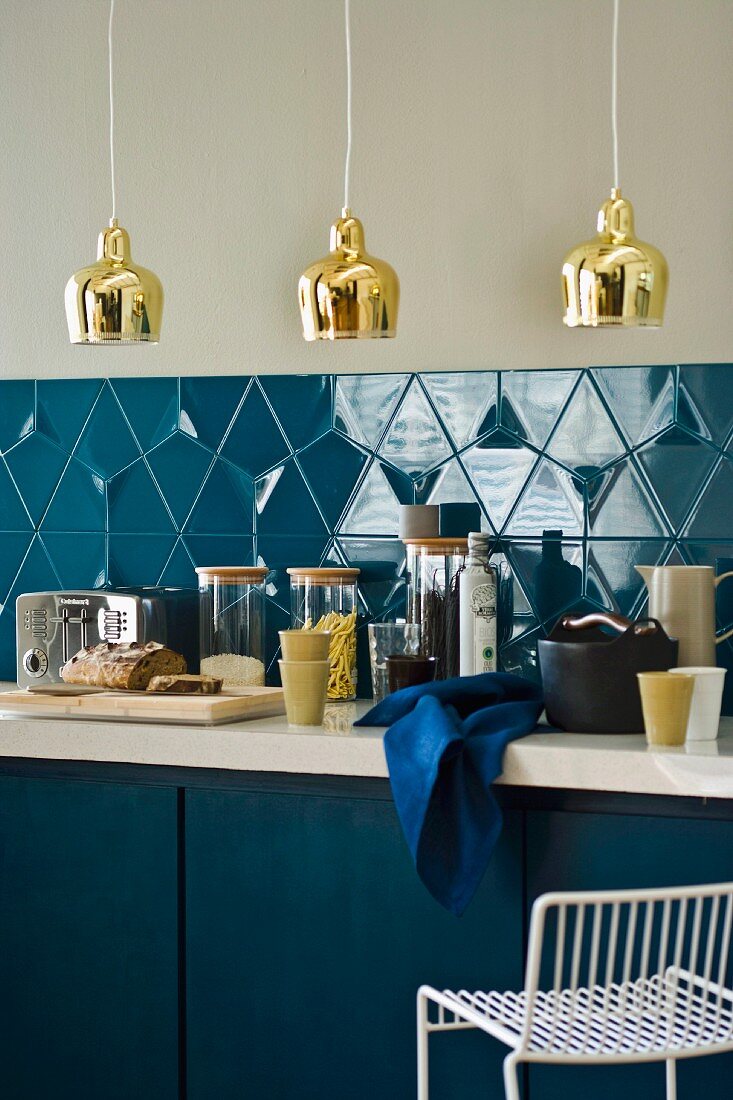 Kitchen counter with blue base units, kitchen utensils in front of blue-tiled splashback & brass pendant lamps
