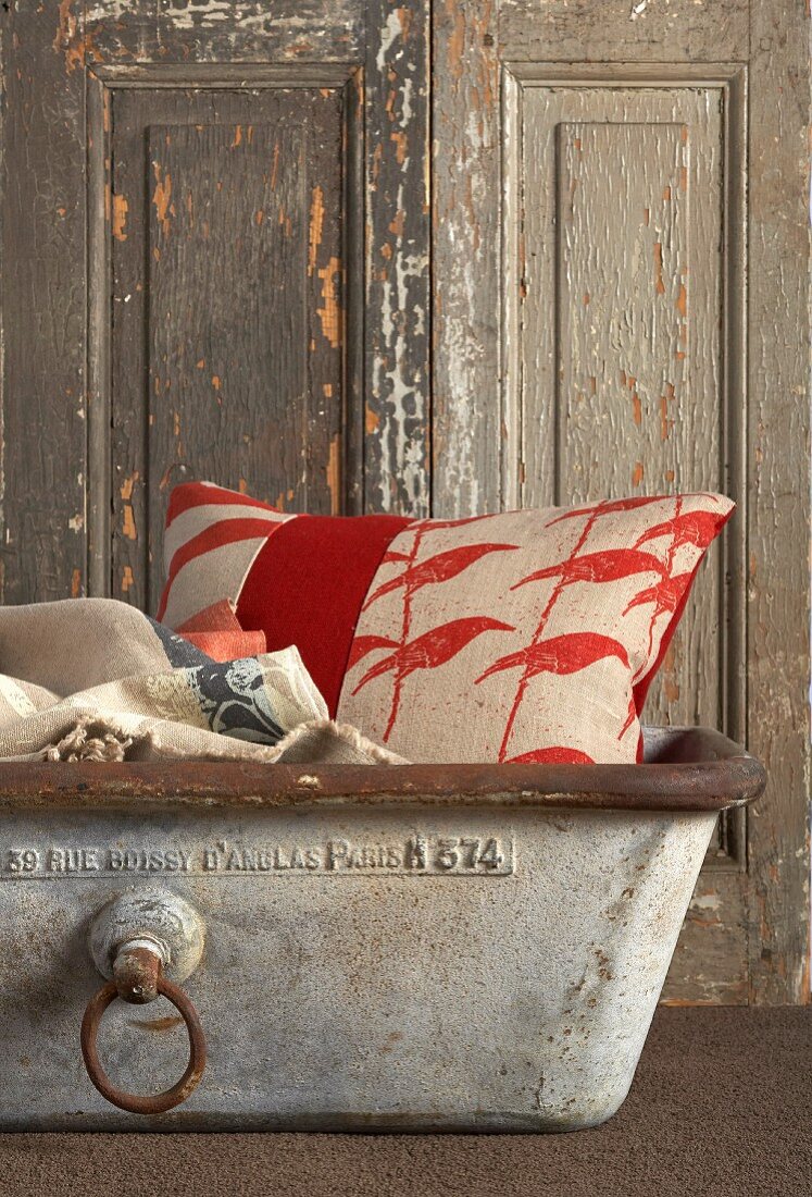 Rot-weiss gemustertes Kissen in Vintage-Metallwanne vor rustikaler Holztür mit abblätternder Farbe