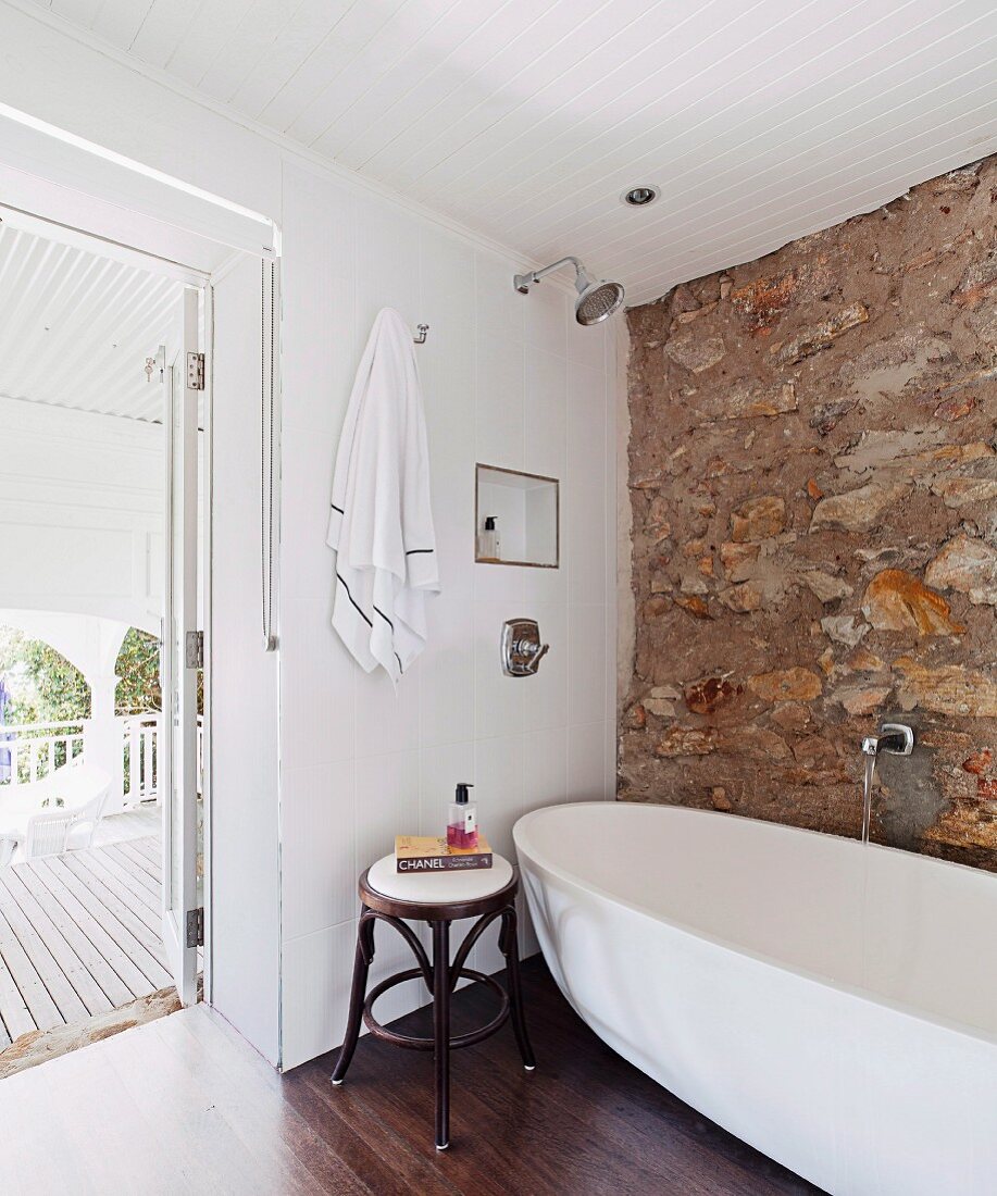 Thonet stool next to modern, free-standing bathtub against stone wall and open terrace door