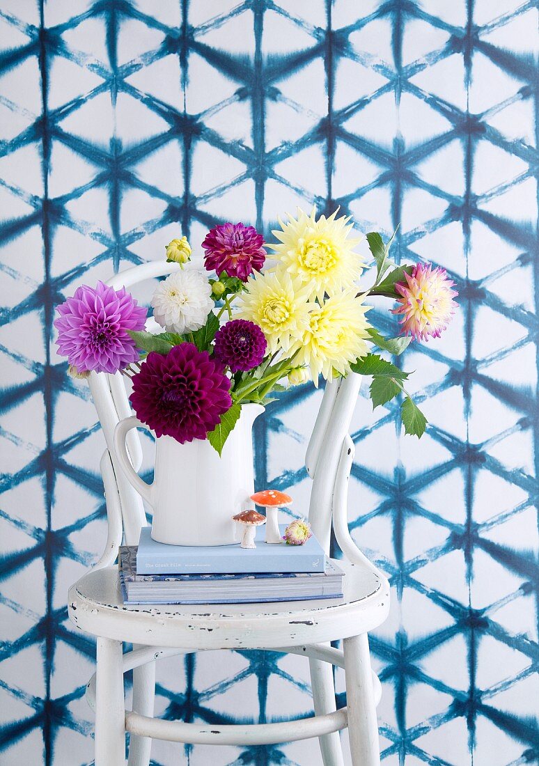 Colourful bouquet of dahlias in white jug and small toadstool ornaments on stacked books on white vintage chair against wall with mesh pattern