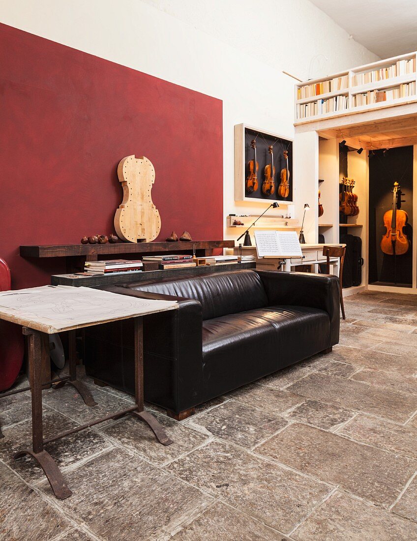 Living area in workshop with string instruments, rustic stone floor, black leather sofa and dark red accent wall panel