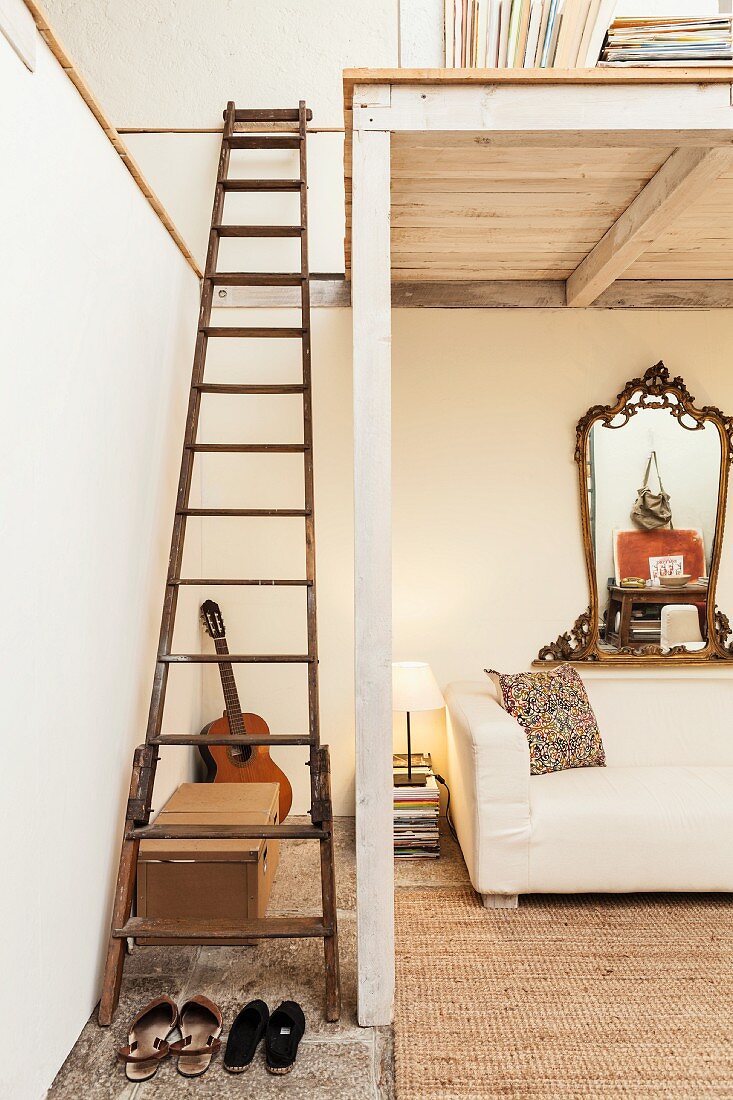 Vintage wooden ladder leading to white-painted wooden mezzanine above ecru sofa and ornate, antique mirror on wall