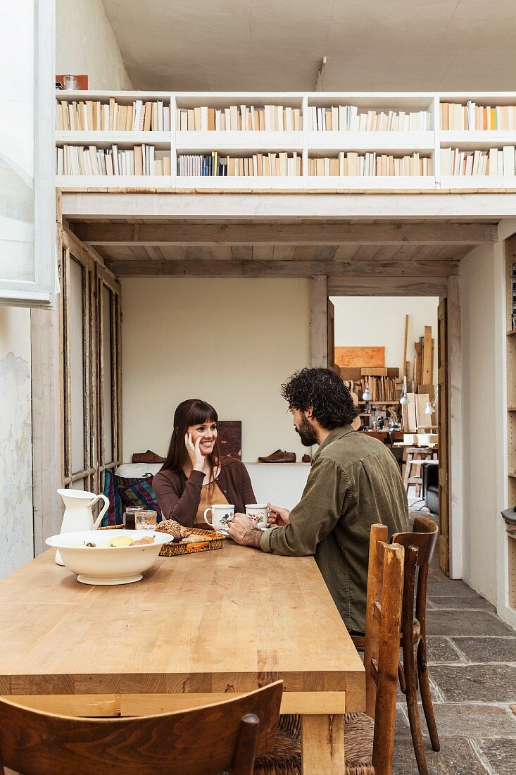 Junges Paar an rustikalem Holztisch, alter grauer Steinboden; Blick in die Werkstatt und auf Bücherregal-Brüstung der Holzgalerie