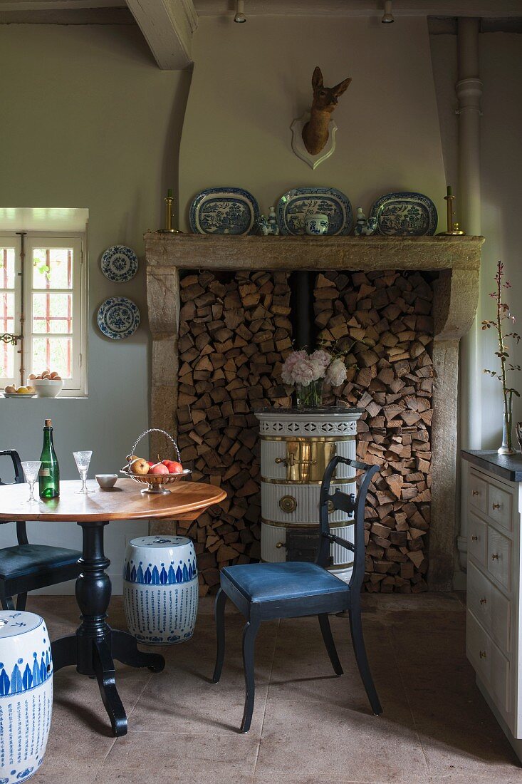 Round table and chair with seat cushion in front of wood-burning stove and stacked firewood in rustic dining room