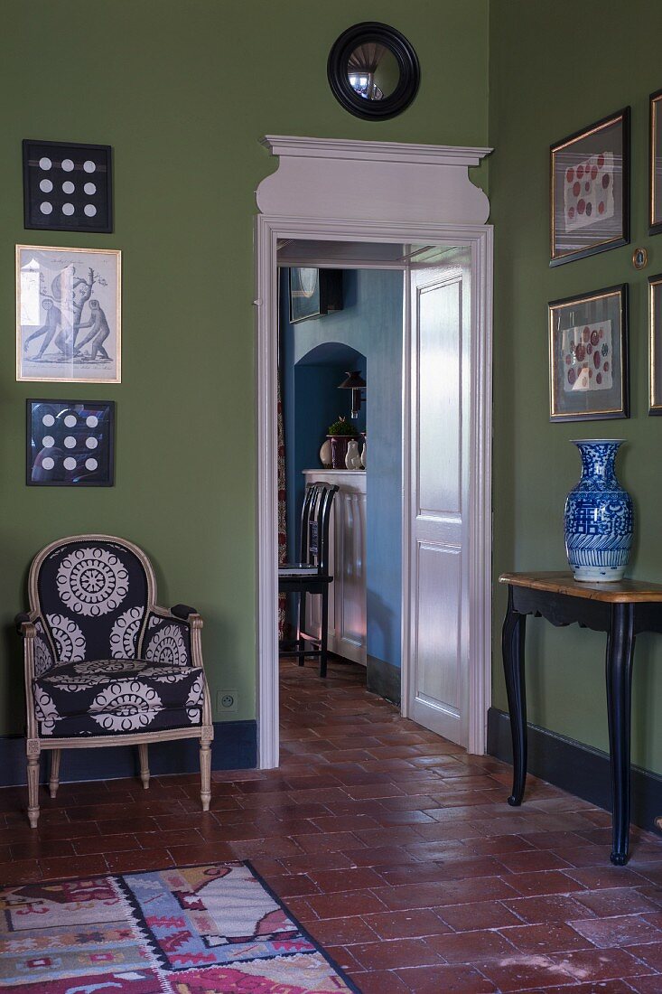 Doorway with white-painted wooden panelling next to armchair and console table below collection of pictures on green-painted wall