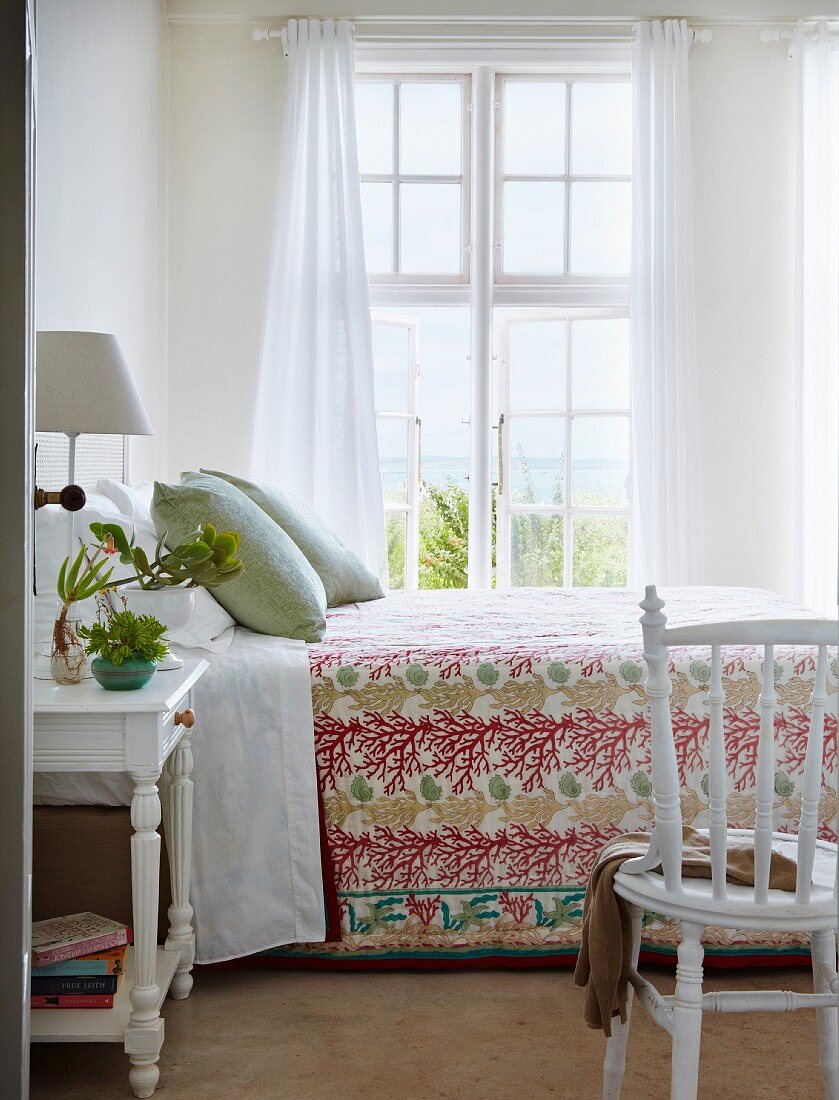 Blick durch offene Tür auf Bett mit Komforthöhe und geblümter Tagesdecke vor Sprossenfenster, luftiger Vorhang