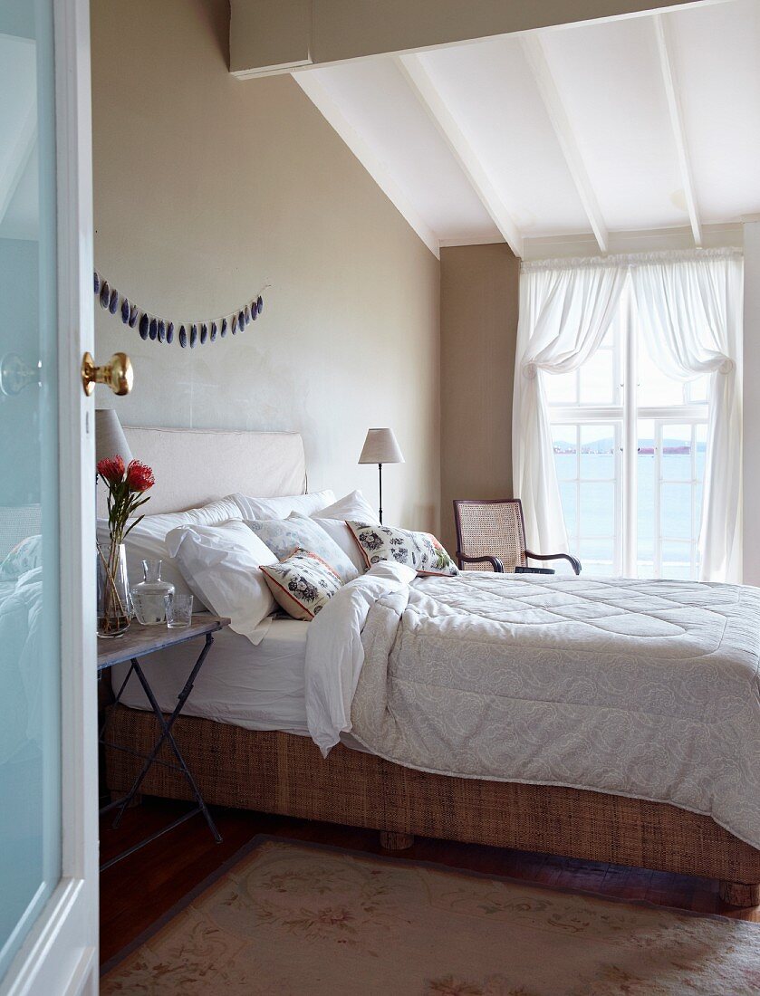 View through open door of double bed at comfortable height in rustic bedroom with taupe walls