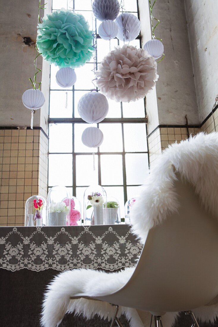 Feminine, romantische Partydekoration mit Pompons und nostalgischer Spitzendecke auf dekoriertem Esstisch in hoher Fabrikhalle mit Industriefenstern