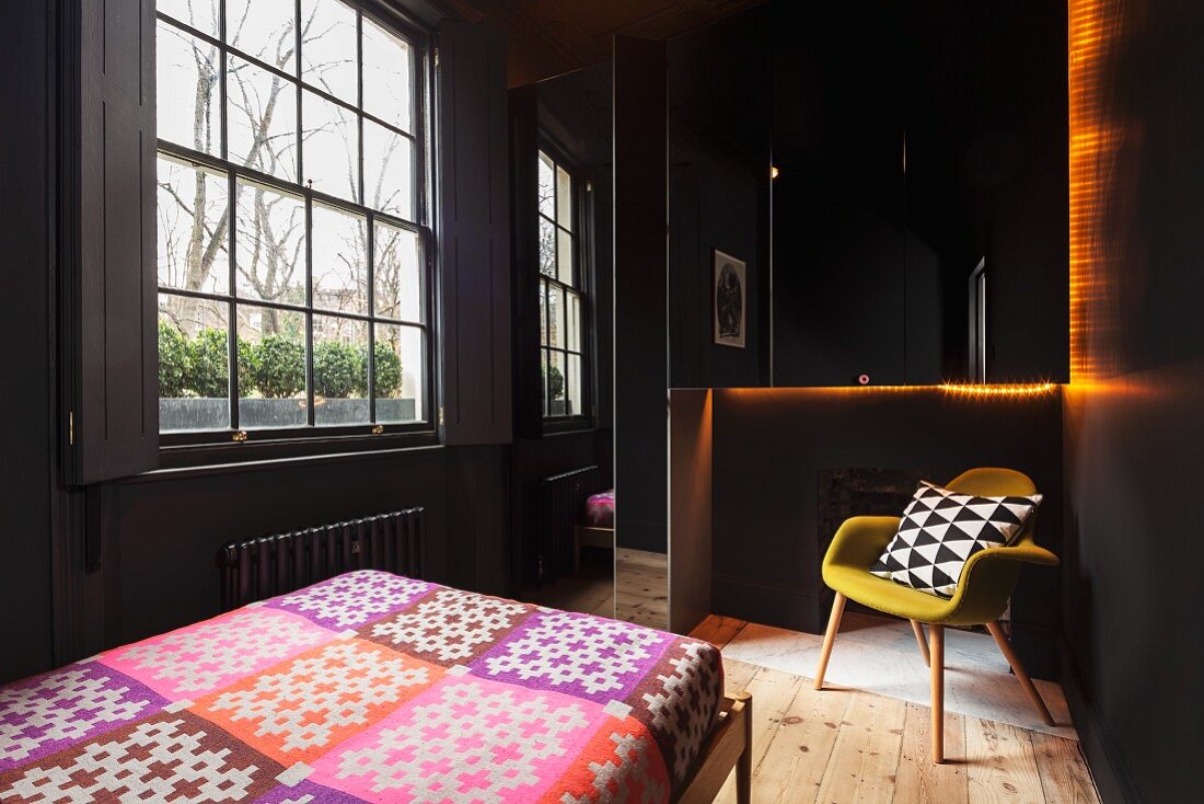 Schwarz gestrichenes Schlafzimmer mit Sprossenfenster, Bett mit bunter Tagesdecke und Retropolsterstuhl auf naturbelassenem Dielenboden