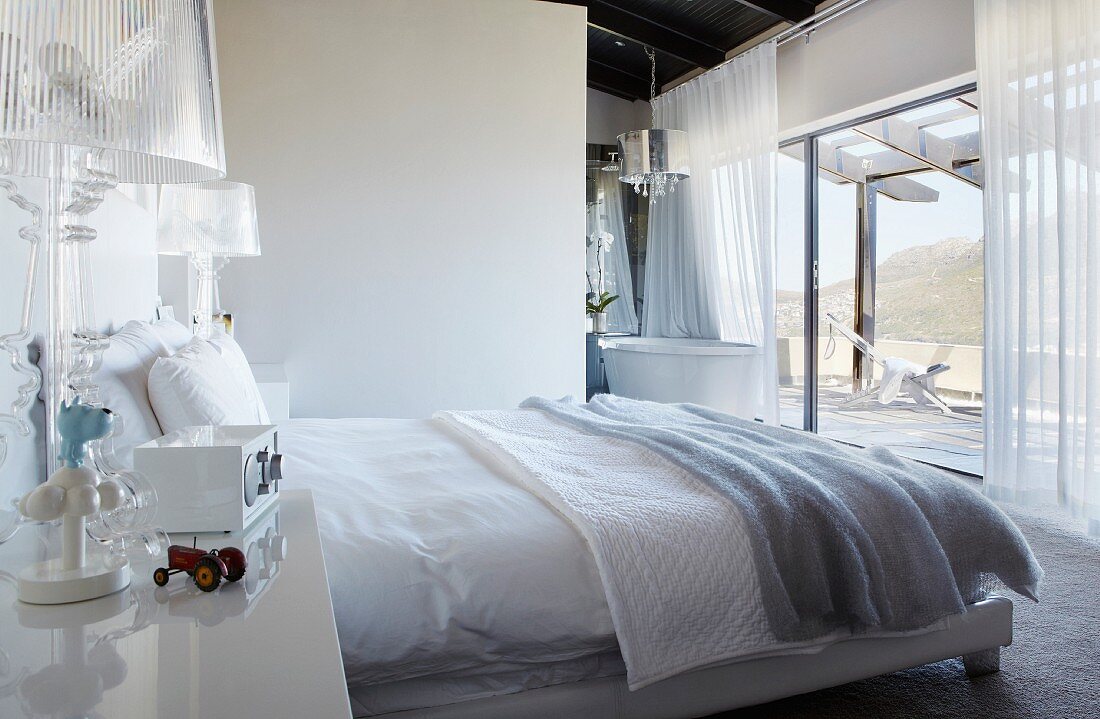 Doppelbett in modernem Schlafzimmer im Hintergrund Bad Ensuite vor raumhohen Terrassenfenstern mit Ausblick