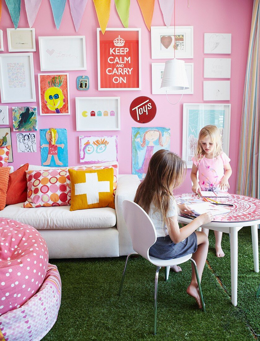 Kinder beim Spielen am runden Tisch, weiße Polstercouch vor rosa Wand mit gerahmter Sammlung von Kinderzeichnungen und grüner Teppich im Rasen-Look