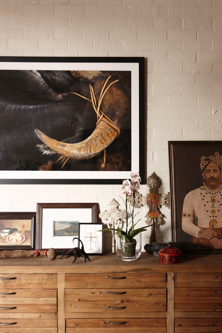 Verschiedene Ölbilder, kunstvolle Kruzifixe mit blühender Orchidee auf Holzsideboard vor weiß gestrichener Ziegelwand mit Gemälde arrangiert