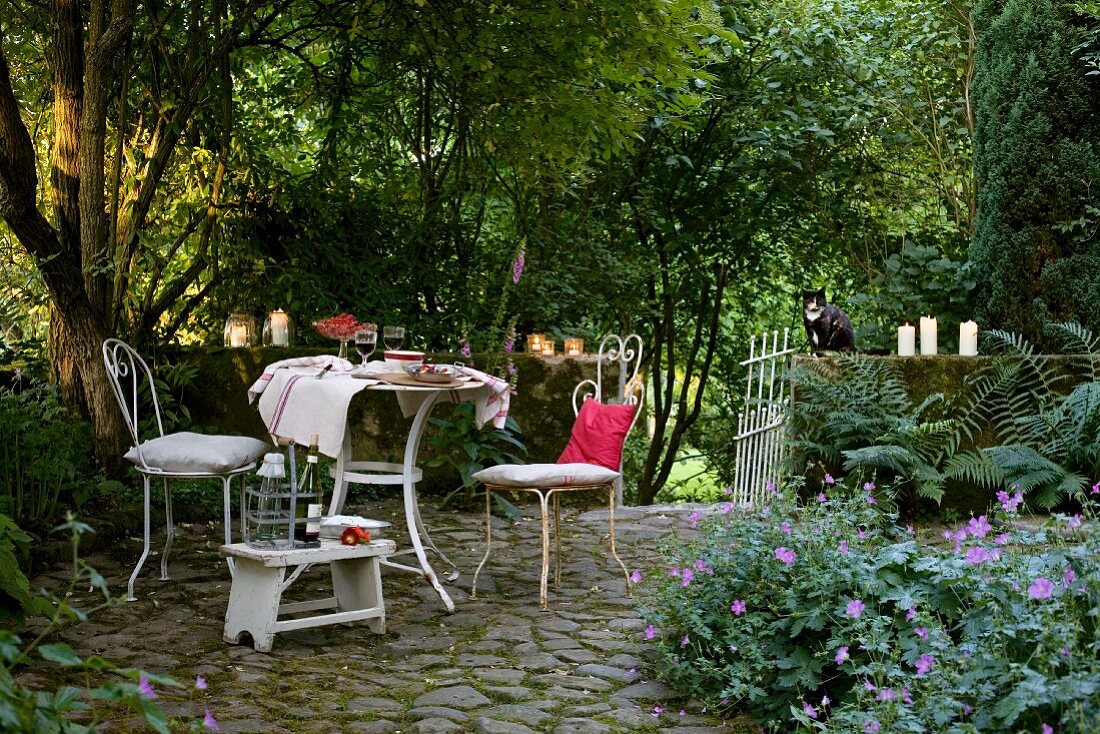 Stimmungsvoller Terrassenplatz - weiße Vintage Metallstühle und Tisch auf Pflasterboden vor Brüstungsmauer mit brennenden Kerzen, dicht bewachsener Garten