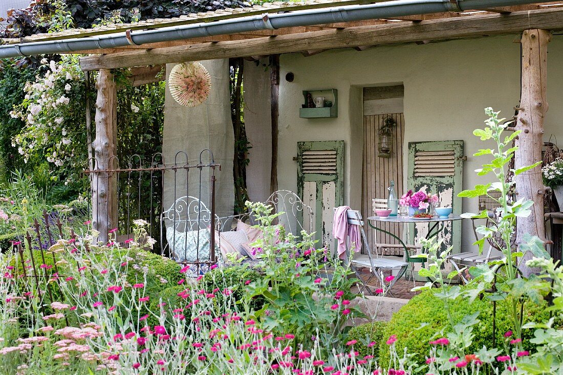 Flowering garden in front of old house with roofed veranda and various vintage metal furnishings