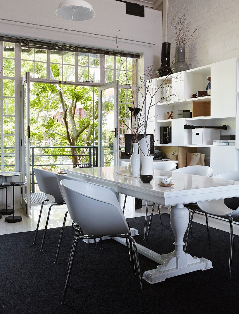 White dining table with turned legs and modern shell chairs on black rug in front of open balcony doors with view of courtyard