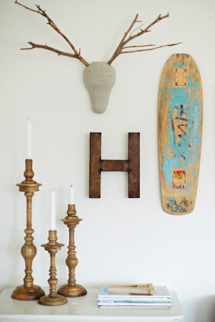 Ausrangiertes Skateboard, Dekobuchstabe-H mit Rostpatina und stilisiertes Ast-Geweih an der Wand über gedrechselten Kerzenleuchtern