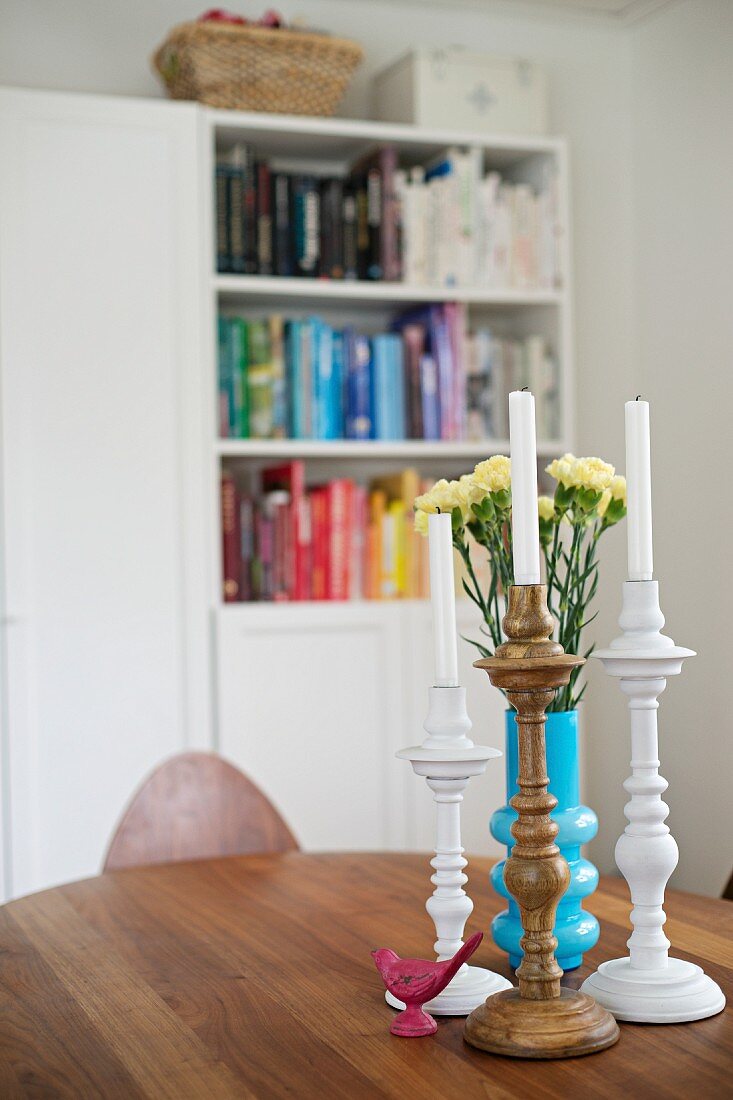Gedrechselte Kerzenständer und blaue Vase mit Nelken auf einem Holztisch; Bücherschrank unscharf im Hintergrund