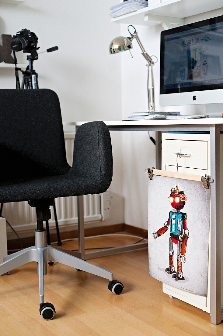Dunkelgrauer Drehstuhl vor Schreibtisch mit Computer, Klammerbügel mit der bunten Illustration eines Roboters an Schubladenschrank